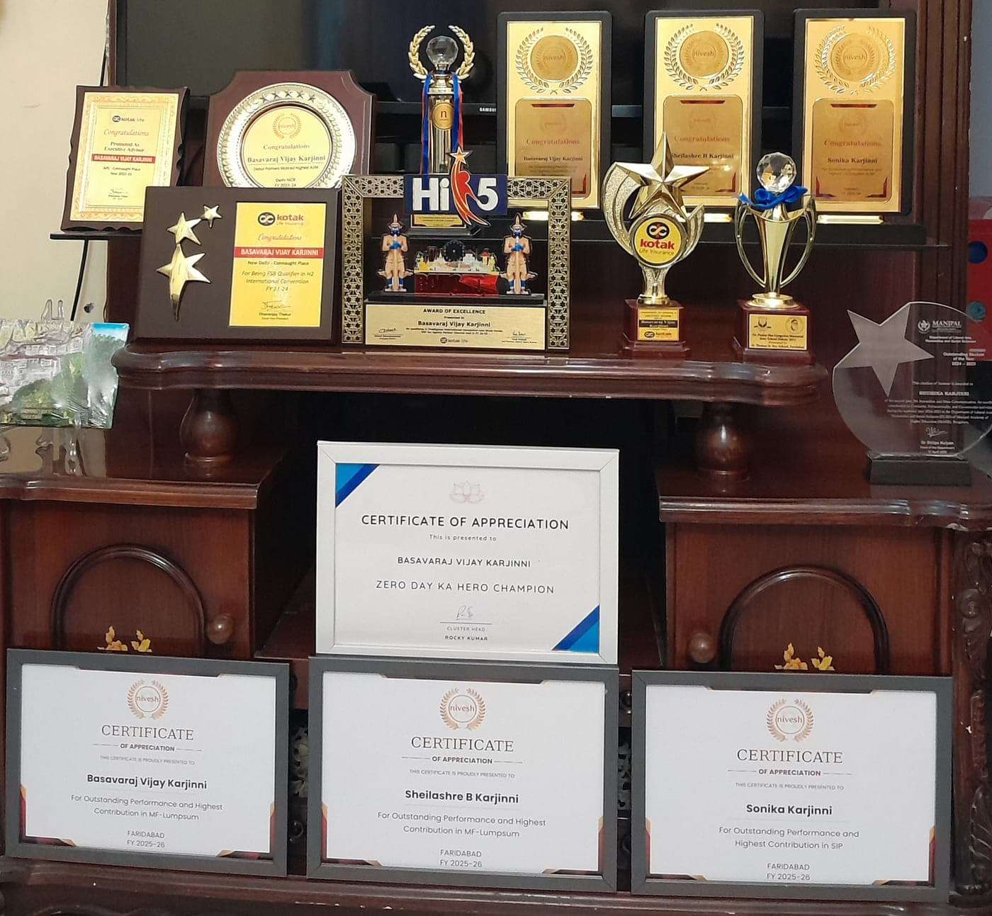 The VijayaRaj award cabinet — Kotak Life and Nivesh plaques, the Hi 5 Bangkok memento, trophies, and FY 2025-26 Nivesh certificates of appreciation for the Karjinni family.